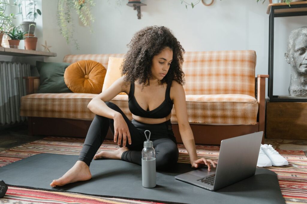 Woman sitting on a yoga mat in activewear, browsing laptop indoors.